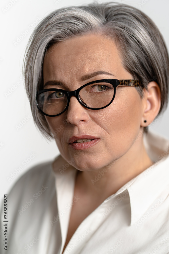 Smart mature woman in white shirt looking thoughtfully at the camera. Pretty Caucasian gray haired model in glasses posing in studio. Close up studio portrait.