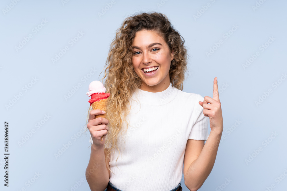 Young blonde woman with curly hair holding a cornet ice cream isolated on blue background intending to realizes the solution while lifting a finger up Young blonde woman with curly hair holding a cornet ice cream isolated on blue background intending to realizes the solution while lifting a finger up