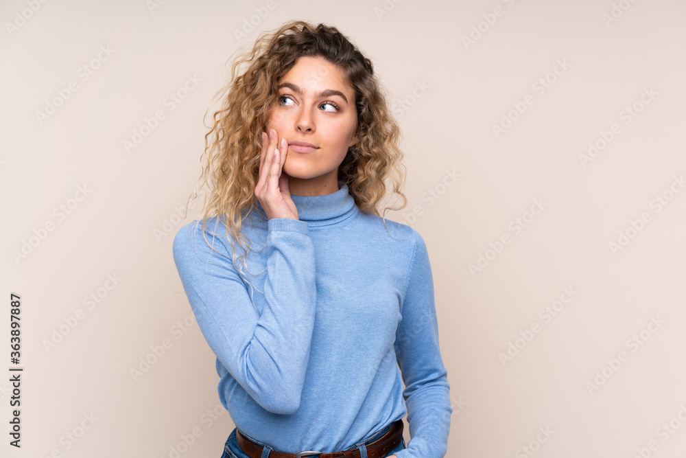 Young blonde woman with curly hair wearing a turtleneck sweater isolated on beige background thinking an idea while looking up Young blonde woman with curly hair wearing a turtleneck sweater isolated on beige background thinking an idea while looking up