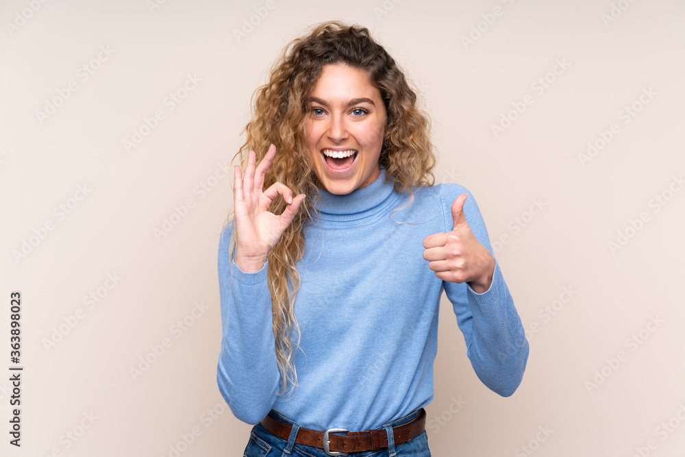 Young blonde woman with curly hair wearing a turtleneck sweater isolated on beige background showing ok sign and thumb up gesture Young blonde woman with curly hair wearing a turtleneck sweater isolated on beige background showing ok sign and thumb up gesture