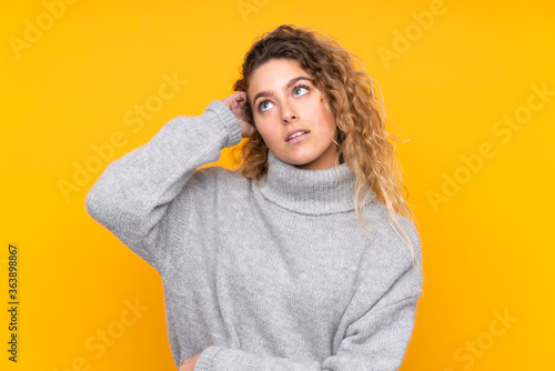 Young blonde woman with curly hair wearing a turtleneck sweater isolated on yellow background having doubts © luismolinero