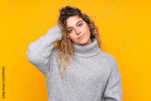 Young blonde woman with curly hair wearing a turtleneck sweater isolated on yellow background with an expression of frustration and not understanding © luismolinero