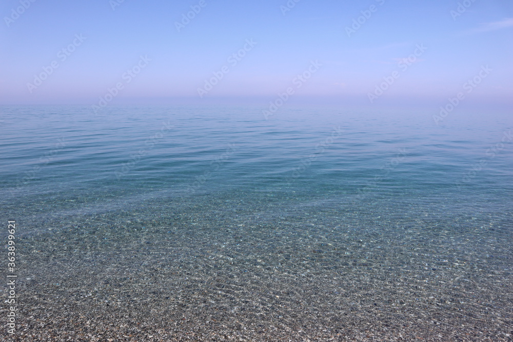 Seascape calm blue sea with transparent water, through which sea pebbles are visible at the bottom on a hot summer day on a pebble beach, blue sky with soft pink haze on the horizon