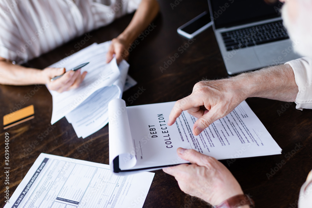 Cropped view of senior man pointing at paper with debt collection lettering near gadgets and wife at home Cropped view of senior man pointing at paper with debt collection lettering near gadgets and wife at home