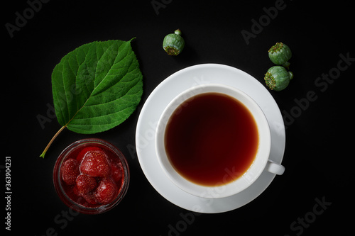 White cup with tea, strawberry jam, poppy boxes and hydrangea leaf on dark background, top view, still life