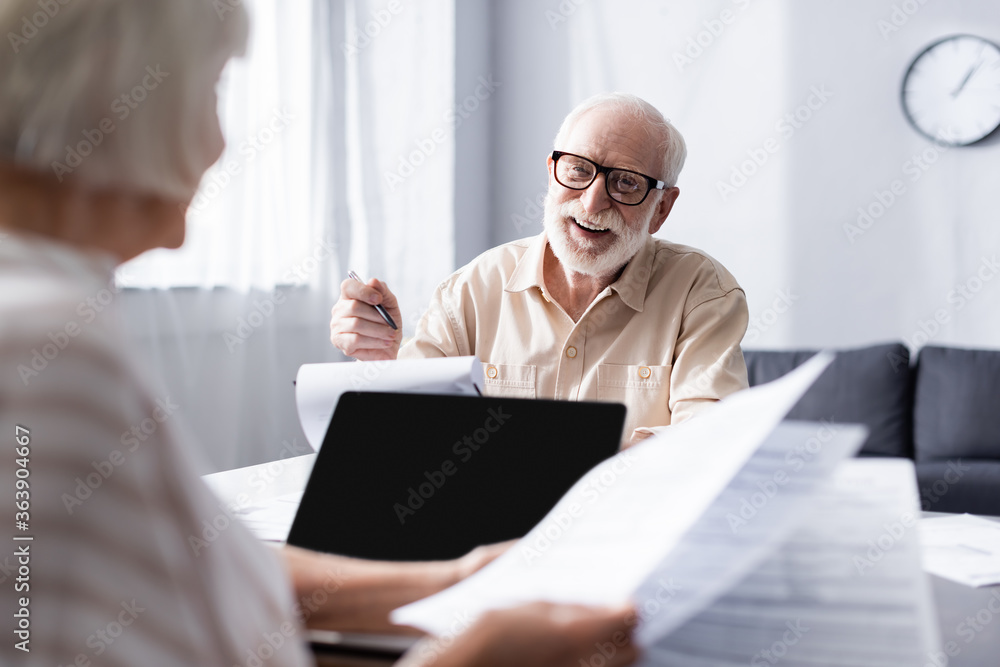 Selective focus of smiling man looking at wife with papers near laptop at home Selective focus of smiling man looking at wife with papers near laptop at home