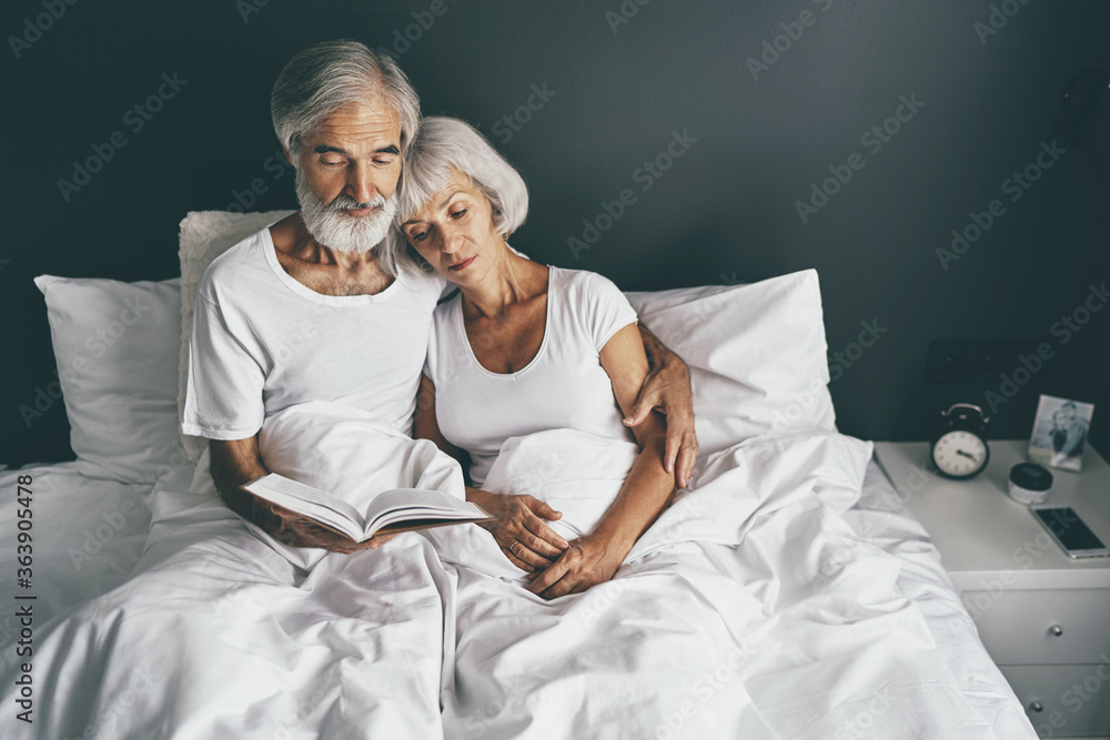 Happy family. Cozy home. Beautiful senior woman and her husband reading book together in bedroom. Happy family. Cozy home. Beautiful senior woman and her husband reading book together in bedroom.