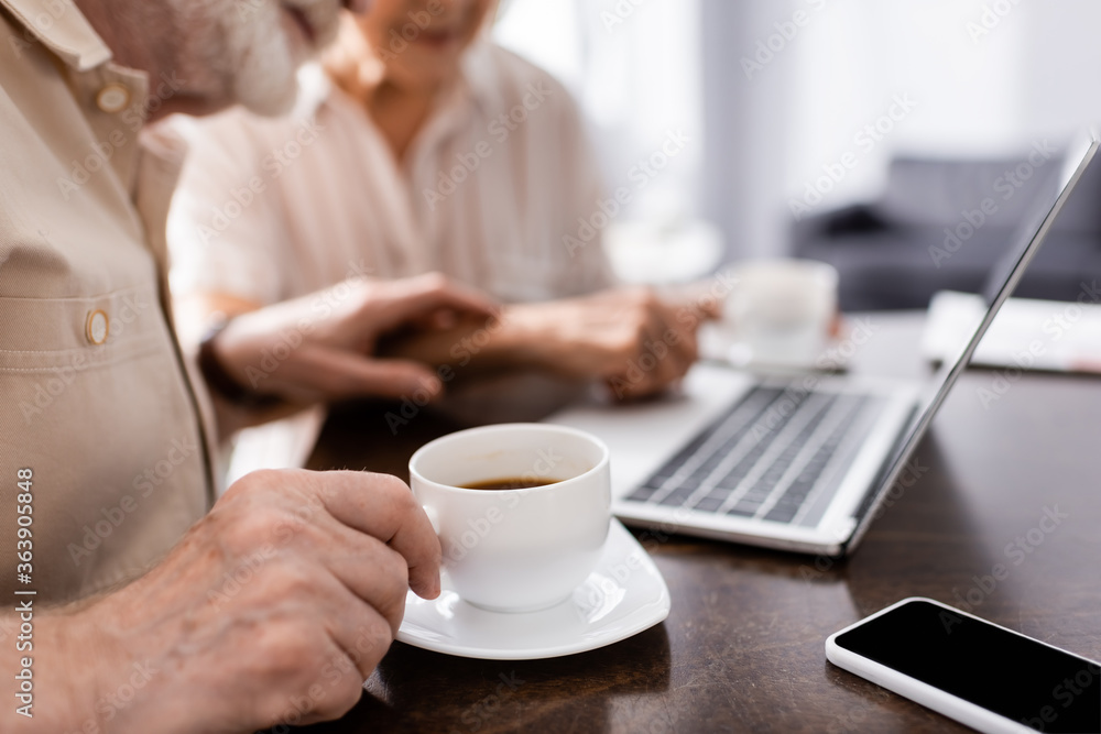 Cropped view of senior man holding cup of coffee near digital devices and wife at home