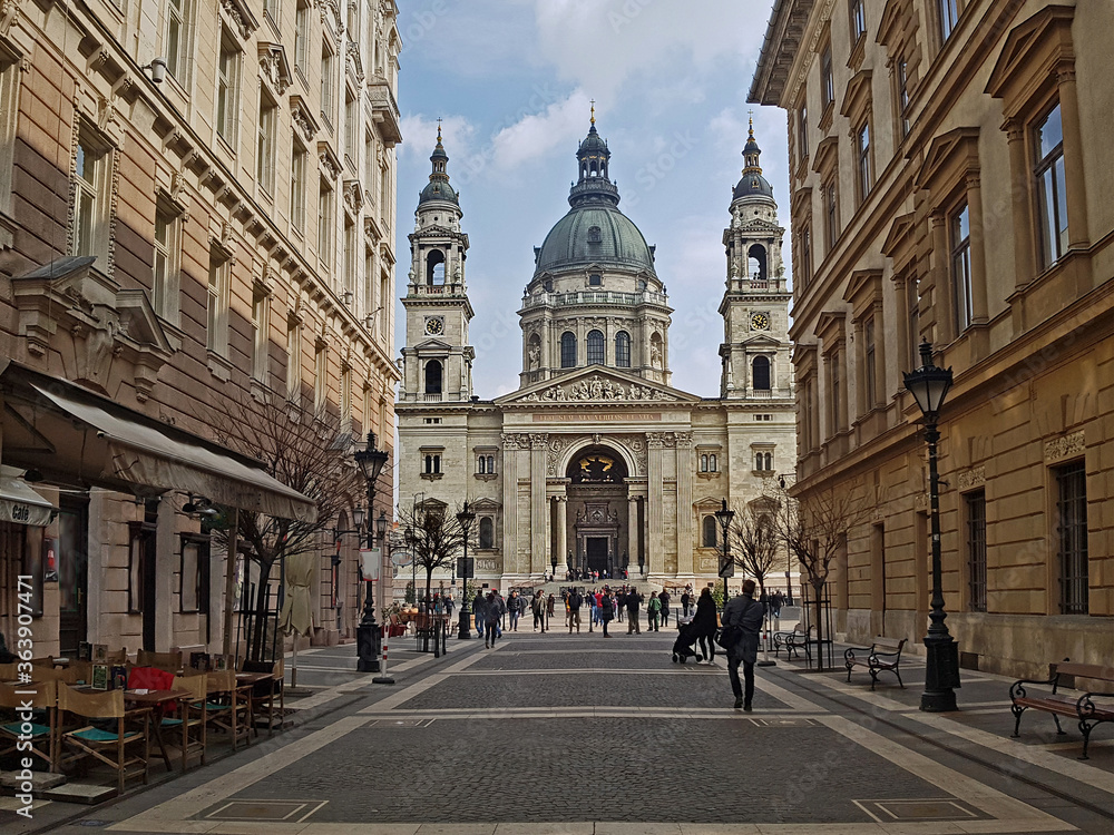 A view on the Roman Catholic St. Stephen's Basilica, seen from the romantical shopping street in front of it, in Budapest, Hungary. The sun lights the church up nicely. Tourists wander over the square