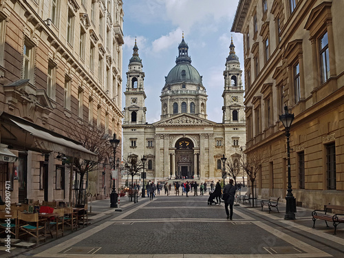 A view on the Roman Catholic St. Stephen's Basilica, seen from the romantical shopping street in front of it, in Budapest, Hungary. The sun lights the church up nicely. Tourists wander over the square