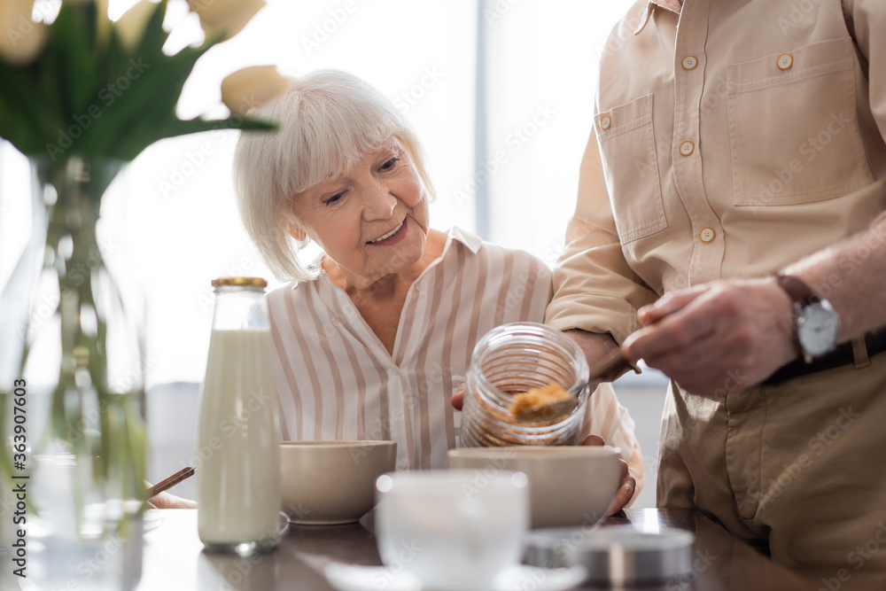 Selective focus of smiling senior woman sitting near man pouring cereals in bowl during breakfast Selective focus of smiling senior woman sitting near man pouring cereals in bowl during breakfast