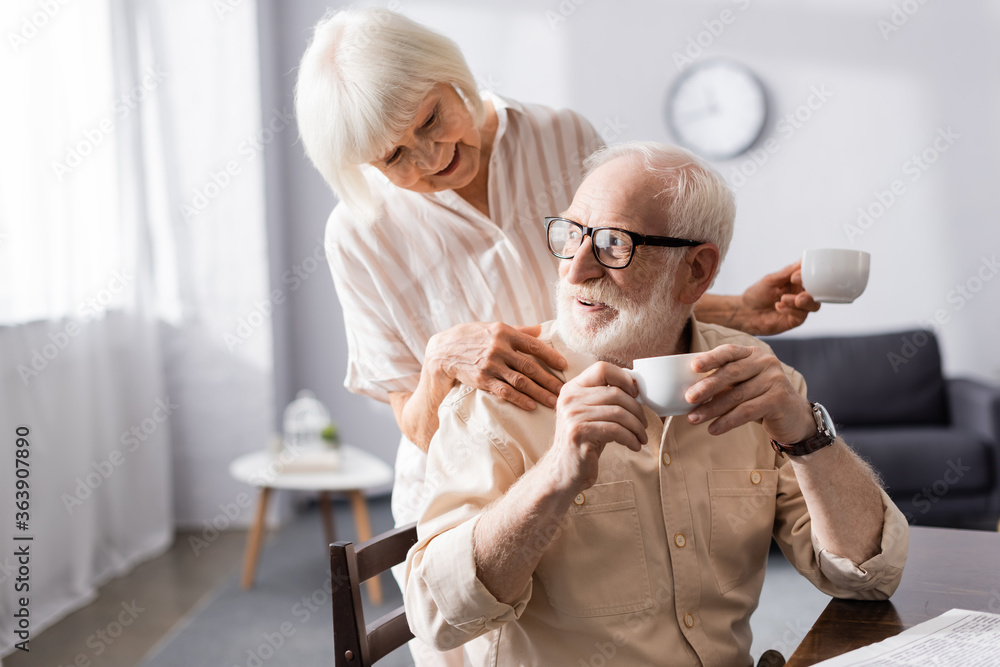 Smiling senior woman holding cup of coffee and embracing husband during breakfast at home Smiling senior woman holding cup of coffee and embracing husband during breakfast at home