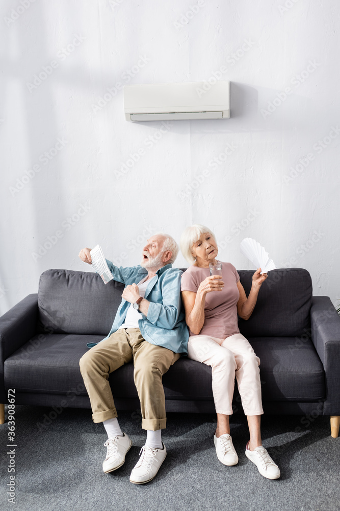 Tired senior couple with glass of water, newspaper and fan feeling hot at home Tired senior couple with glass of water, newspaper and fan feeling hot at home