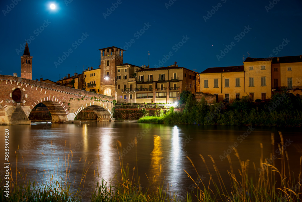 Night photo with moon in sky view along Adige river with view of Ponte della Pietra with tower and bell tower Santa Anastasia, city of Verona, Italy.