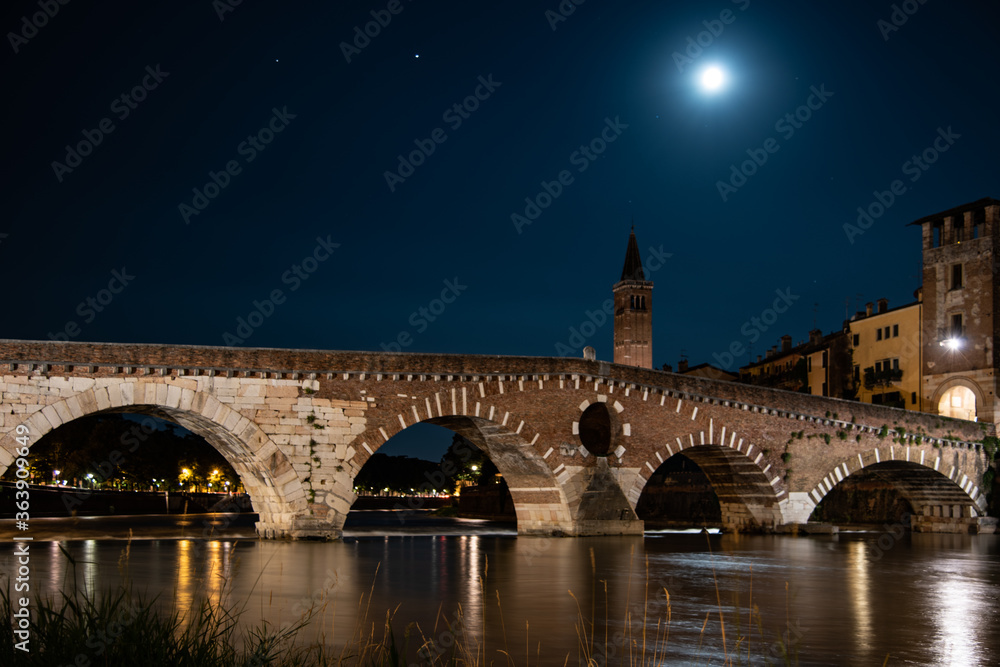 Night photo with moon in sky view along Adige river with view of Ponte della Pietra and campanile Santa Anastasia, city of Verona, Italy.