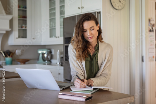Woman taking notes while using laptop in the kitchen