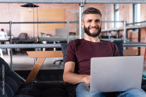 cheerful businessman sitting on sofa and using laptop in office