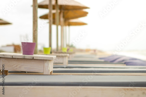 Focus on the cup on the wooden table next to the sun lounger on the beach bar. The beach is empty and there is no one.
