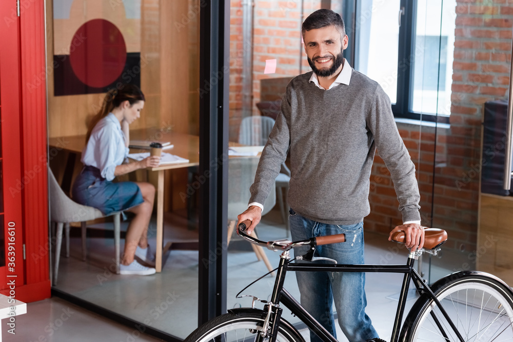 selective focus of cheerful businessman standing with bicycle near woman sitting in office
