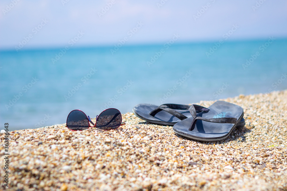 Selective focus on old slippers. Next are sunglasses. They are located on the yellow sand on the beach and it is a beautiful sunny day. Copy space. Selective focus on old slippers. Next are sunglasses. They are located on the yellow sand on the beach and it is a beautiful sunny day. Copy space.