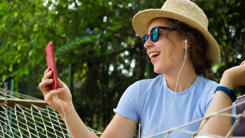 Excited young woman in headphones using mobile phone relaxing on hammock,video calling, sharing data on social media,making selfie,listening music,podcast,blogging.Girl in summer hat and sunglasses