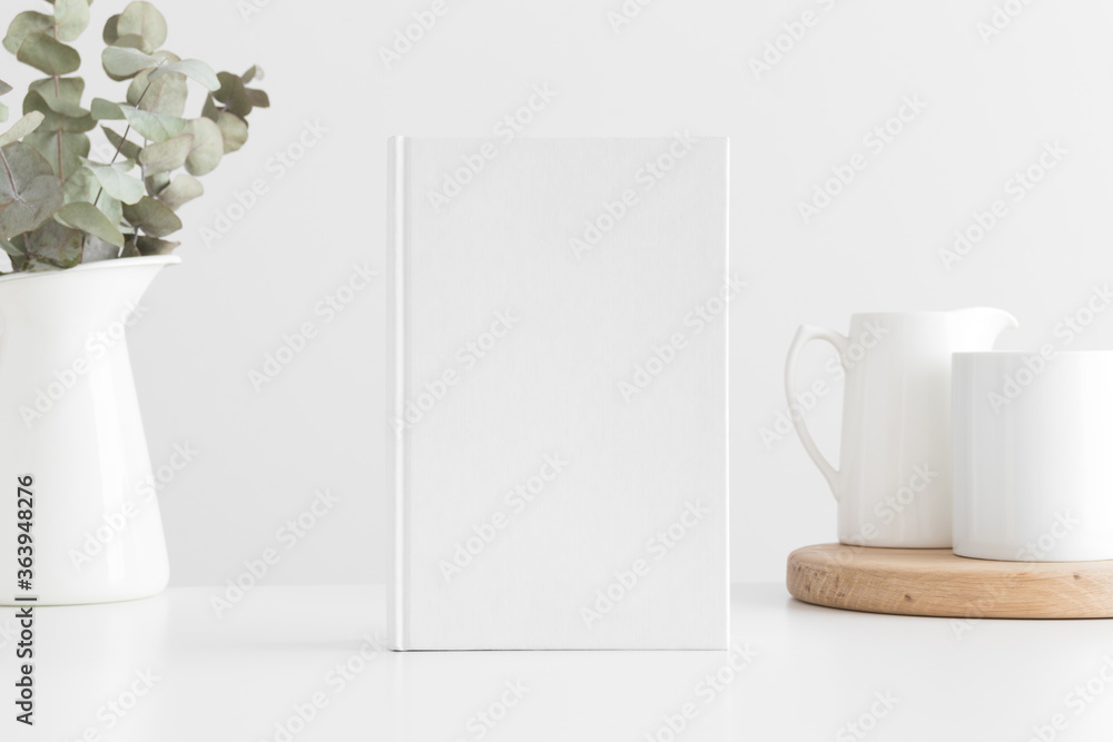 White book mockup with a eucalyptus in a pot and a mug on a white table.