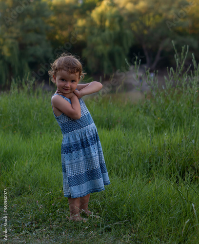 Cute little girl posing with a smile for the camera. She folded her hands under her chin. Dressed in a blue dress. Against a background of green grass.
