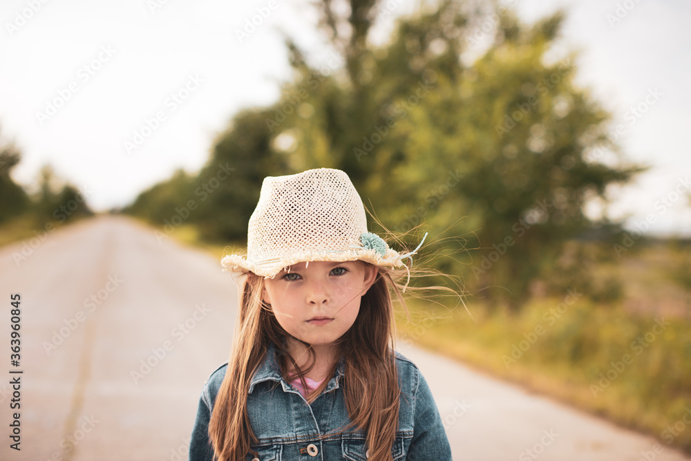 Portrait of a beautiful little girl in a straw hat and denim jacket against the backdrop of a rural road. High quality photo