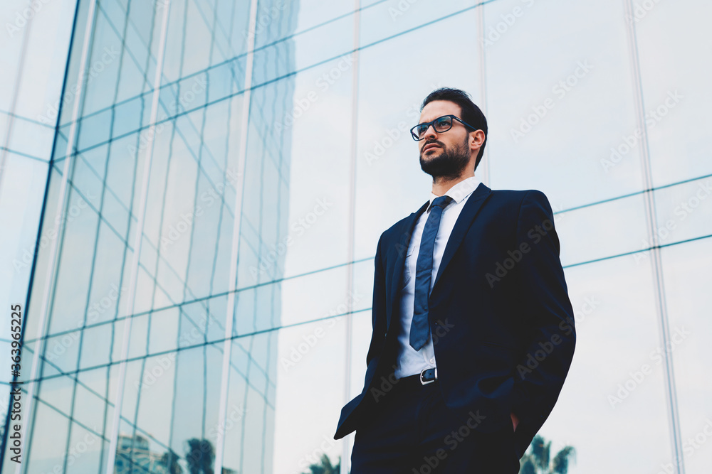 Confident business man in glasses standing against office building with hands in the pockets Confident business man in glasses standing against office building with hands in the pockets