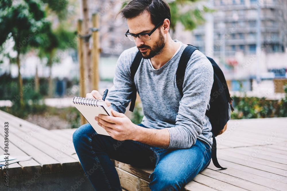 Obraz premium Skilled handsome male international student of journalism faculty in eyewear making homework task creating essay inspiring of good weather outdoors spending leisure during break in university campus