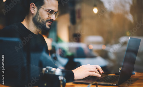Bearded entrepreneur male in glasses dressed in black sweater reading news checking email and chatting with friends in social network using wireless internet connection at wooden table in coffee shop