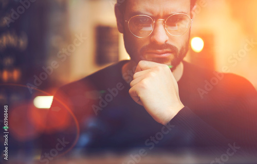 Cropped close up image of pensive bearded entrepreneur in optical spectacles for better views dressed in black sweatshirt.Pondering businessman in trendy eyeglasses thinking on blurred background