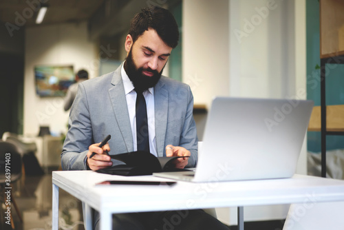 Professional male manager of It company planning working schedule noting details while checking working projects on laptop computer, owner of business corporation creating plans on new startup