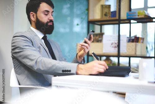 Handsome executive manager dressed in elegant formal outfit booking tickets online on website via smartphone noting important information in diary, businessman writing down contact of colleague