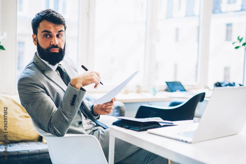 Confident serious entrepreneur with beard sitting with documentation thinking on new strategy plannning working at laptop computer in office while looking away near copy space area for advertsing text Confident serious entrepreneur with beard sitting with documentation thinking on new strategy plannning working at laptop computer in office while looking away near copy space area for advertsing text