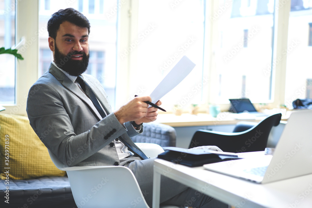 Portrait of smiling bearded executive manager of trading corporation checking financial report sitting in coworking office, businessman satisfied with working achievements and successful startup