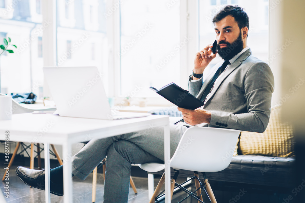 Portrait of serious bearded economist having mobile conversation with colleague on telephone device while checking financial reports sitting at workplace near copy space area for business information