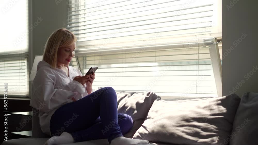 A close-up portrait of a nice woman in a middle-aged white shirt with short blond hair, sitting on a sofa with pillows in the office, holding a phone. A close-up portrait of a nice woman in a middle-aged white shirt with short blond hair, sitting on a sofa with pillows in the office, holding a phone.