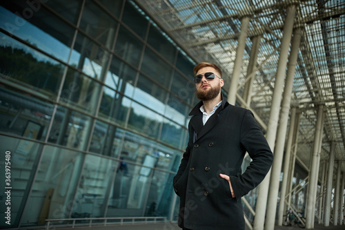 Success. Stylish young bearded man in coat, sunglasses businessman standing near the business center, looking far. With hands pockets, pensive and concentrated. Confident look of