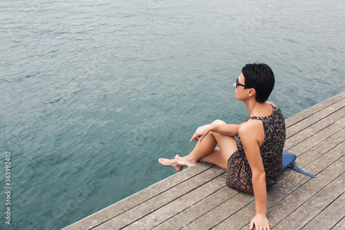 Portrait of a young charming woman enjoying recreation time during her summer weekend  female tourist in sunglasses sitting on a wooden pier against river with copy space area for your text message