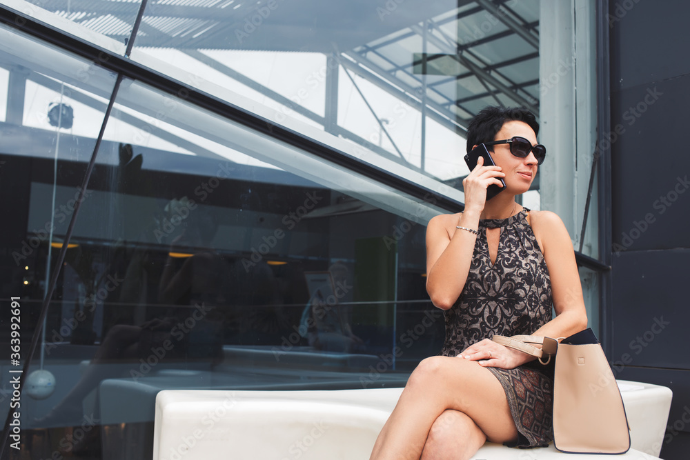 Portrait of wealthy businesswoman in sunglasses using smart phone while looking aside, beautiful female employed speaking on the phone while sitting alone near modern glassy building