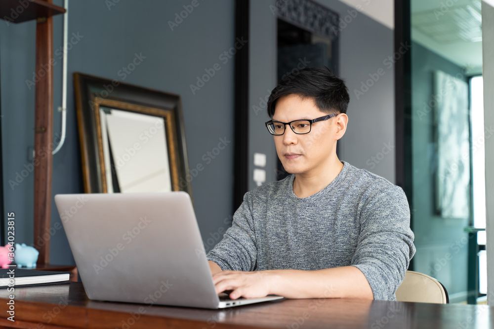 Confident busy Asian male freelancer in eyewear working on laptop computer at moder home. Confident busy Asian male freelancer in eyewear working on laptop computer at moder home.