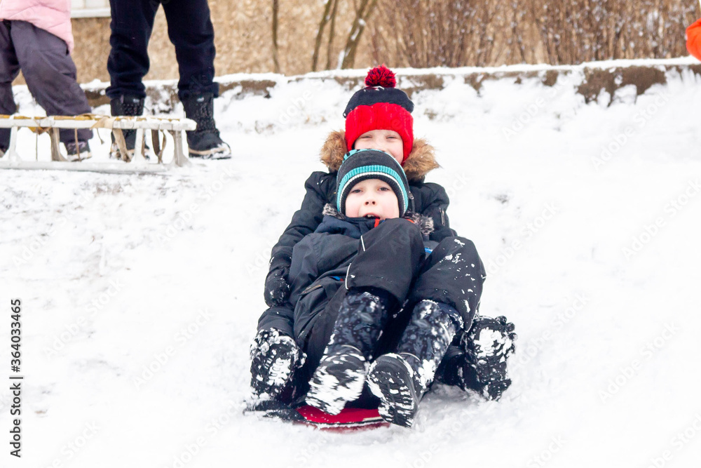 A boy rides from a snow slide on a red cardboard. Fun games in the snow.  Two friends are lying in the snow. Snowy winter in Russia.