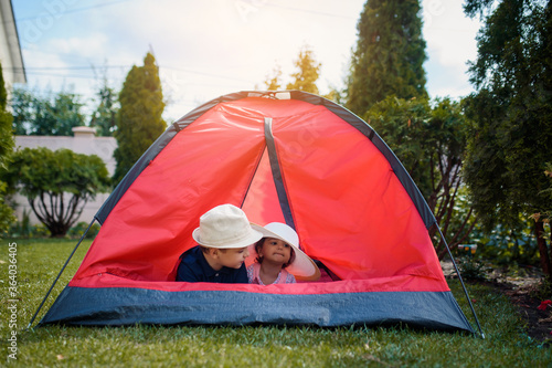 Two happy little kids boy and girl brother and sister are playing in a red camping tent in the home yard on the grass.