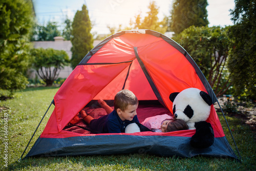 Two happy little kids, boy and girl brother and sister are lying and playing in a red camping tent with their panda bear toy in the home yard on the grass.