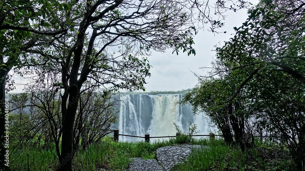 Victoria Falls, Zimbabwe. In the foreground is green grass, trees, a path, a fence in front of the precipice. Further visible powerful streams of water falling in the gorge. Fog above the waterfall. Victoria Falls, Zimbabwe. In the foreground is green grass, trees, a path, a fence in front of the precipice. Further visible powerful streams of water falling in the gorge. Fog above the waterfall.