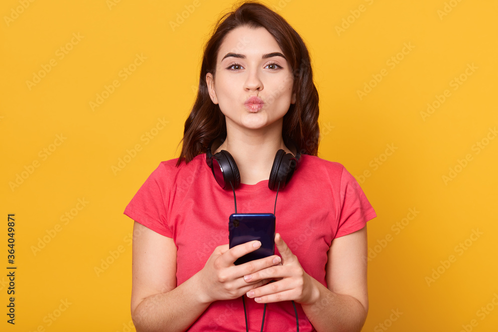 Young attractive girl with phone in hands, showing kiss gesture with her lips, having headphones around neck, standing isolated over yellow background. Young attractive girl with phone in hands, showing kiss gesture with her lips, having headphones around neck, standing isolated over yellow background.