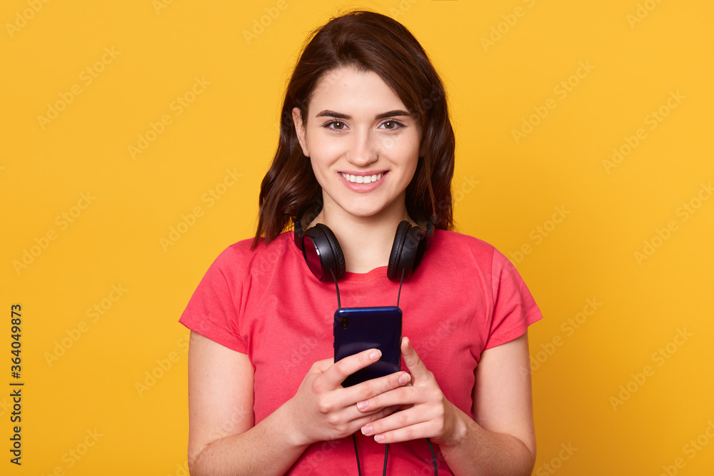 Indoor shot of happy smiling female with dark hair, looking directly at camera, dresses casually, posing isolated over yellow background with phone in hands and has headphones around neck. Indoor shot of happy smiling female with dark hair, looking directly at camera, dresses casually, posing isolated over yellow background with phone in hands and has headphones around neck.