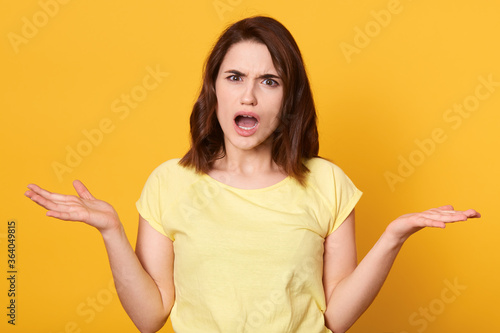Closeup of puzzled unhappy woman isolated over yellow background showing negative emotions, looking at camera with opened mouth, throwing her palms up.