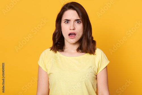 Astonished woman with pleasant appearance and dark beautiful hair, looking directly at camera, keeping mouth widely opened, expressive shock, looks surprised, wearing yellow t shirt. © sementsova321